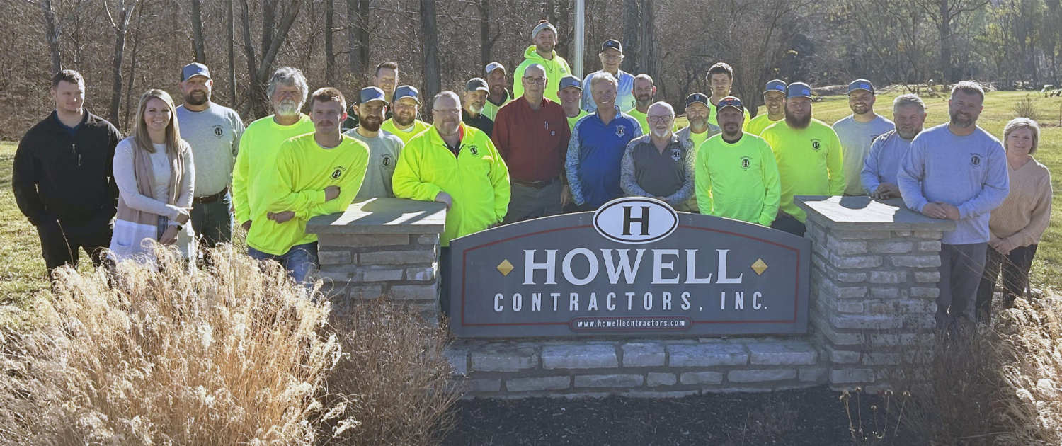 The Howell Contractors team of professionals stands proudly next to the company sign on a sunny autumn day.